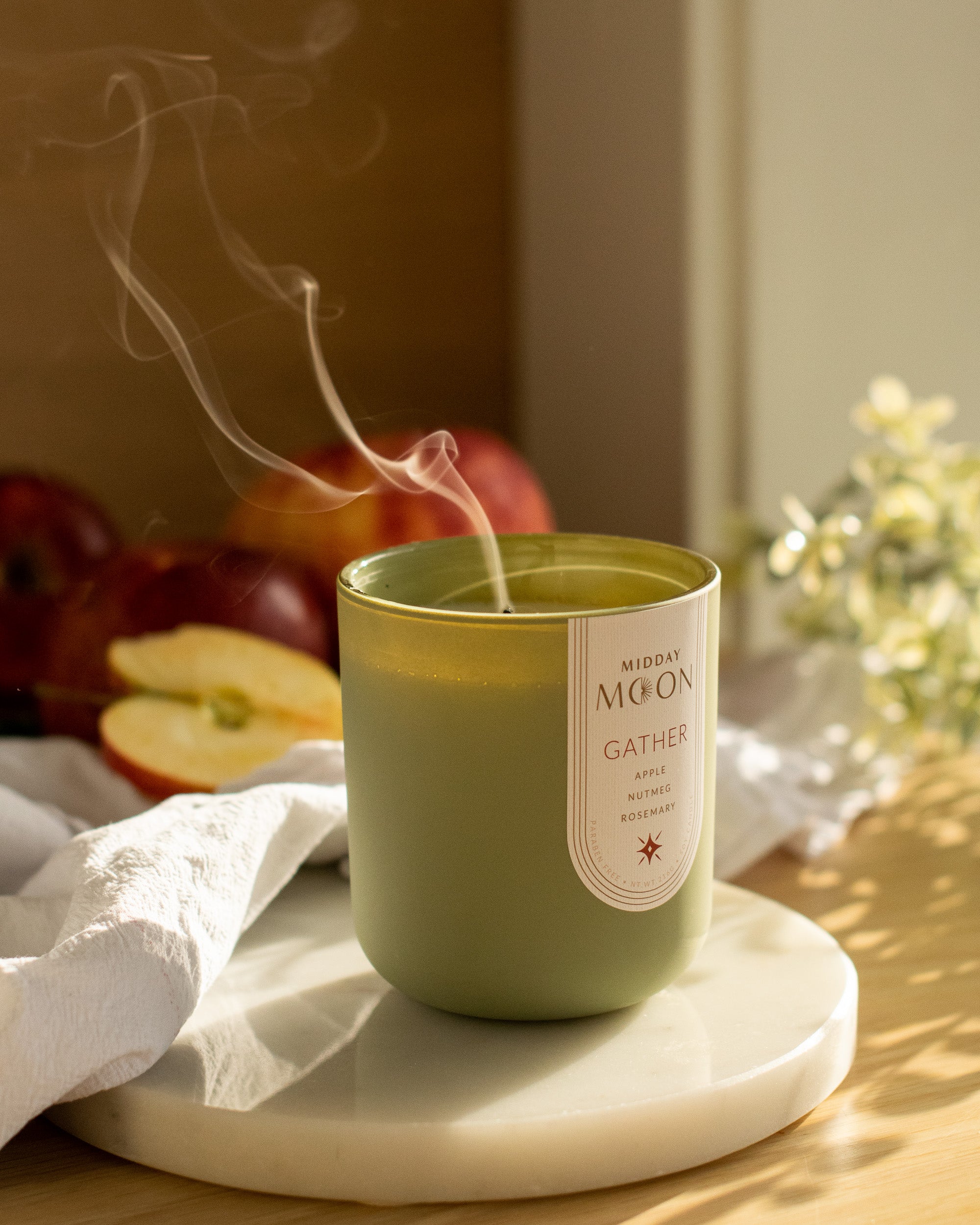 Green candle labeled 'Midday Moon' on a marble surface with apples and flowers in the background