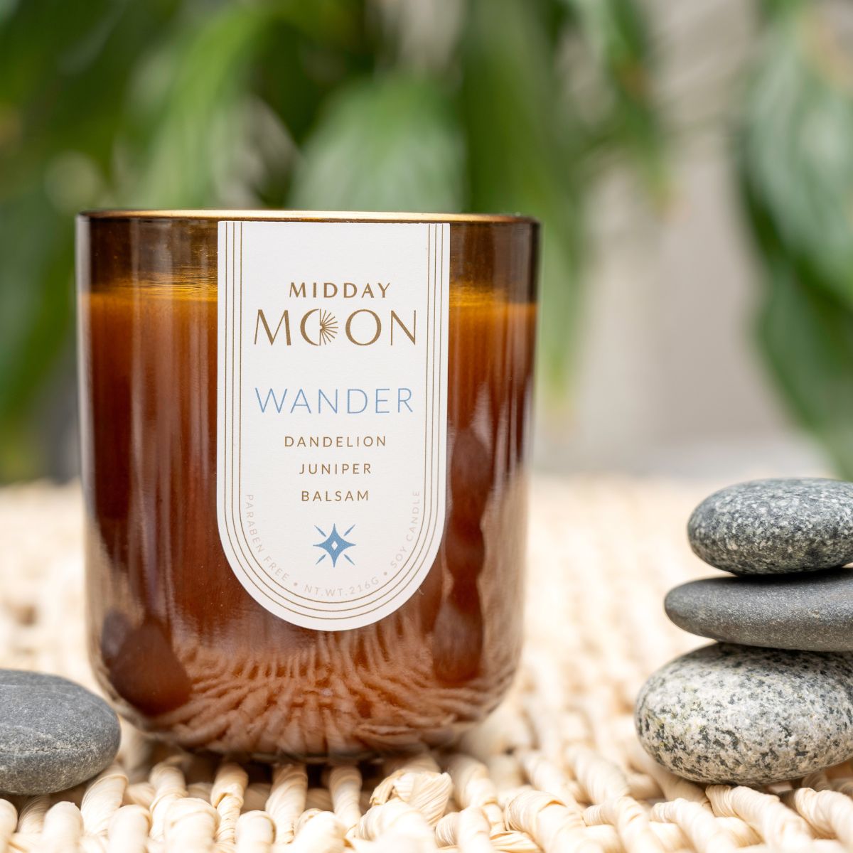 closeup of a soy candle in an amber jar. Wander candle scented with dandelion, juniper, and balsam. Staged with smooth river rocks.