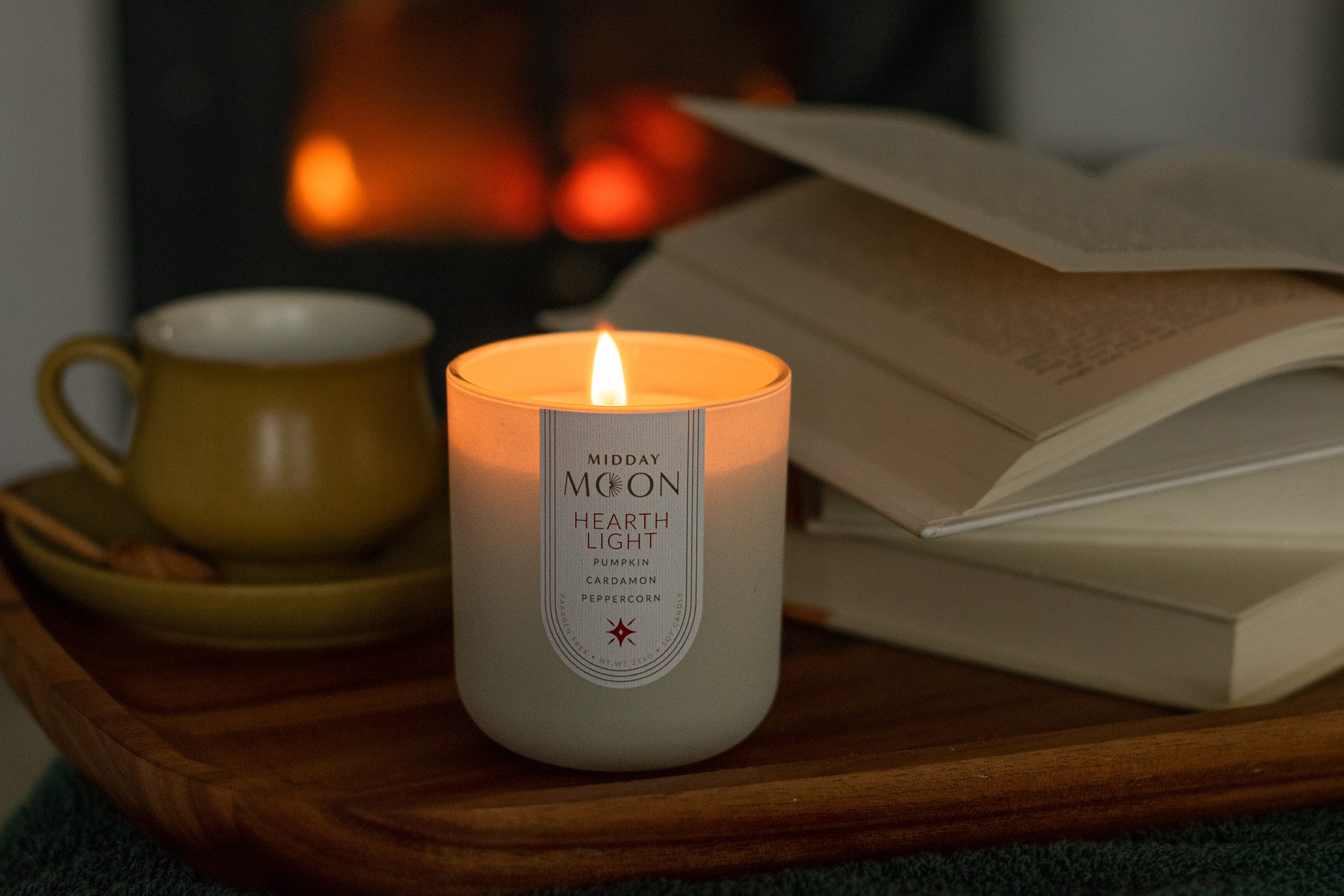 Hearthlight candle lit in the foreground with a mug, and book on a wooden tray in front of a fireplace in the background.