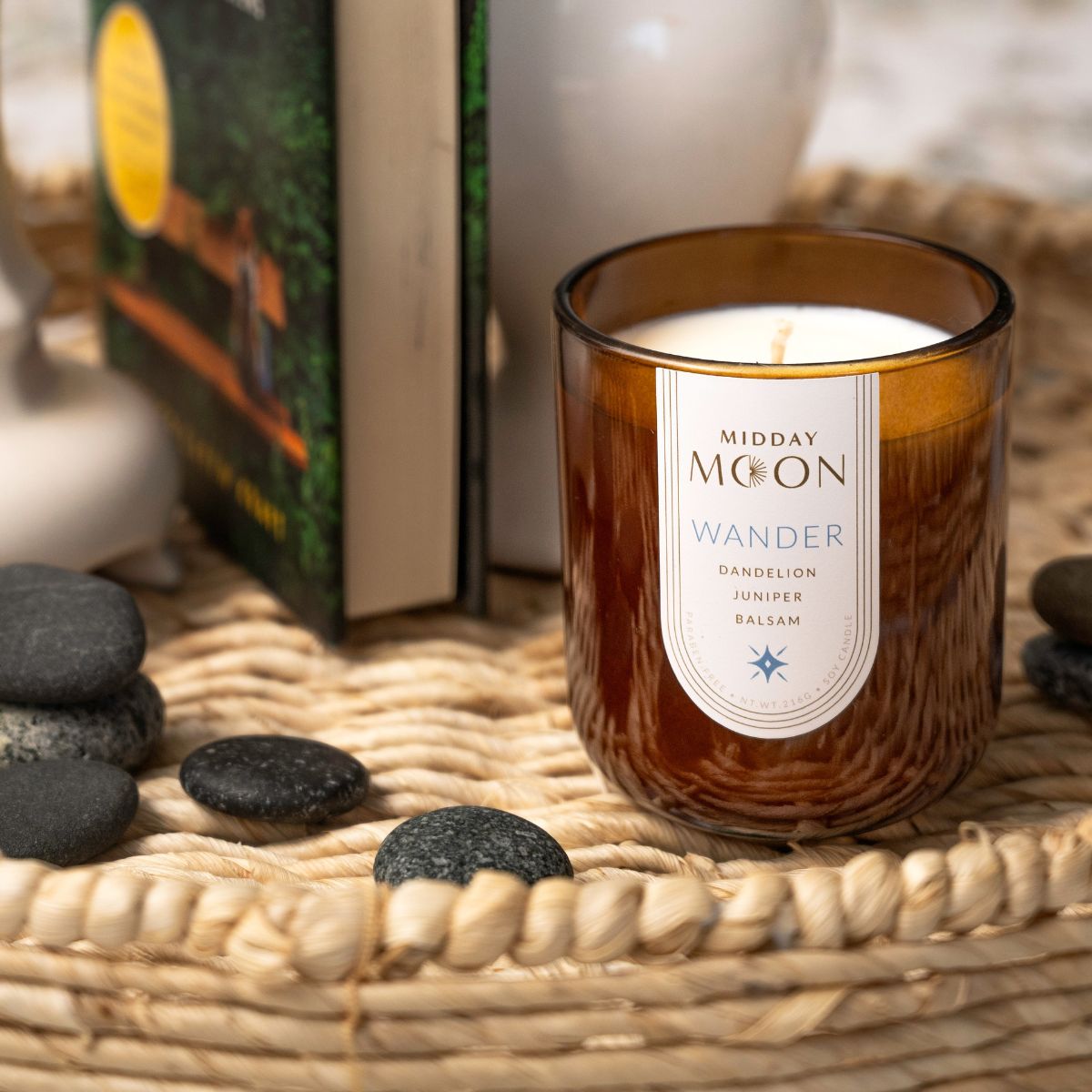 Scented soy candle in an amber jar. Midday moon candle scented with dandelion, juniper, and balsam on a rattan tray.